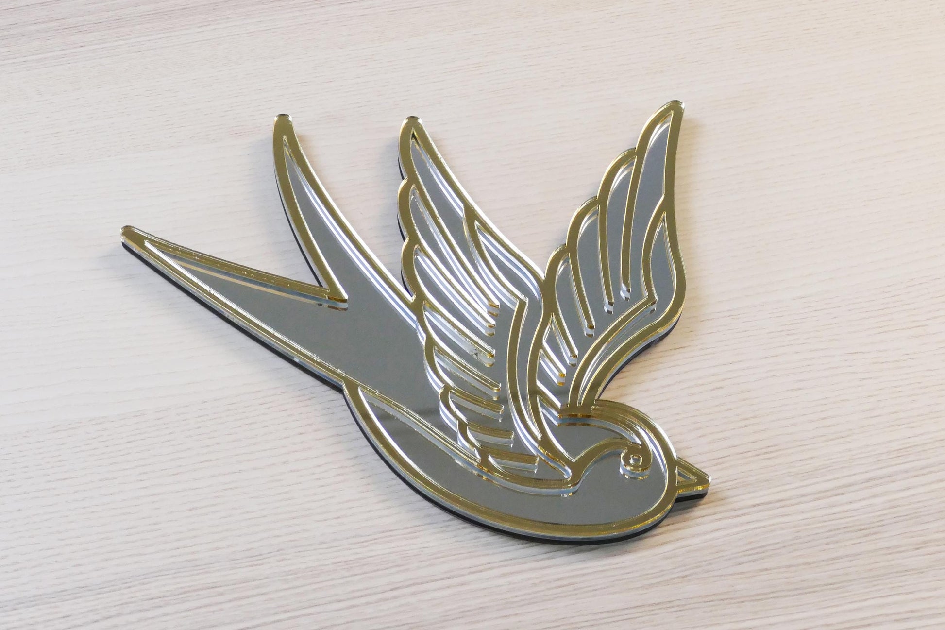 Gouden en zilveren acryl spiegel in tattoo-stijl van een zwaluw, hangend aan een muur.