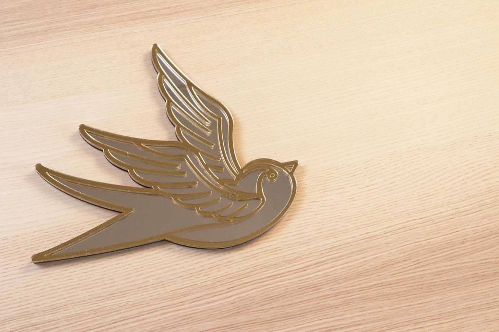 Gouden en zilveren acryl spiegel in tattoo-stijl van een zwaluw, hangend aan een muur. 
