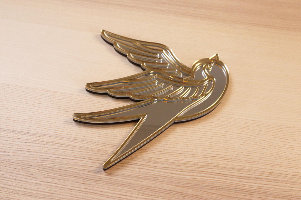 Gouden en zilveren acryl spiegel in tattoo-stijl van een zwaluw, hangend aan een muur.